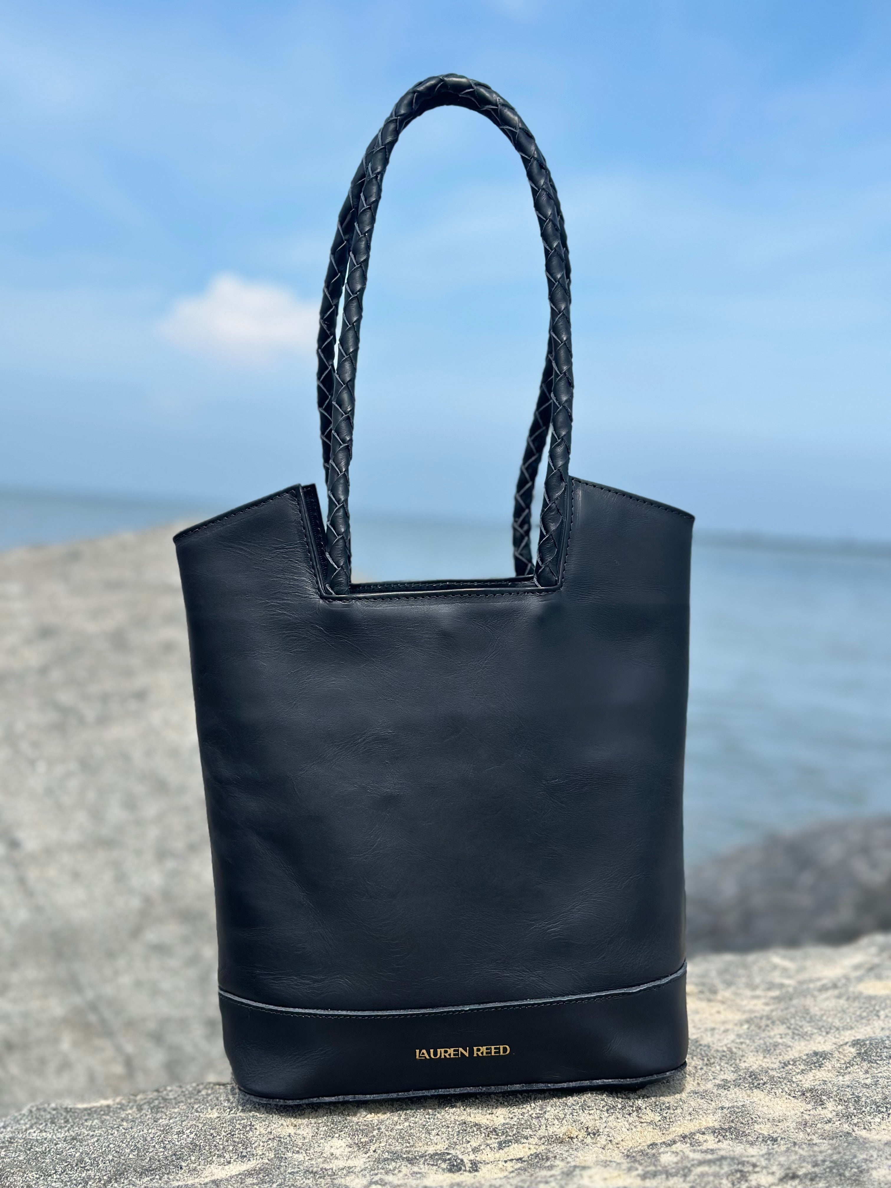 Black tote bag with braided handle on a stone surface with ocean view