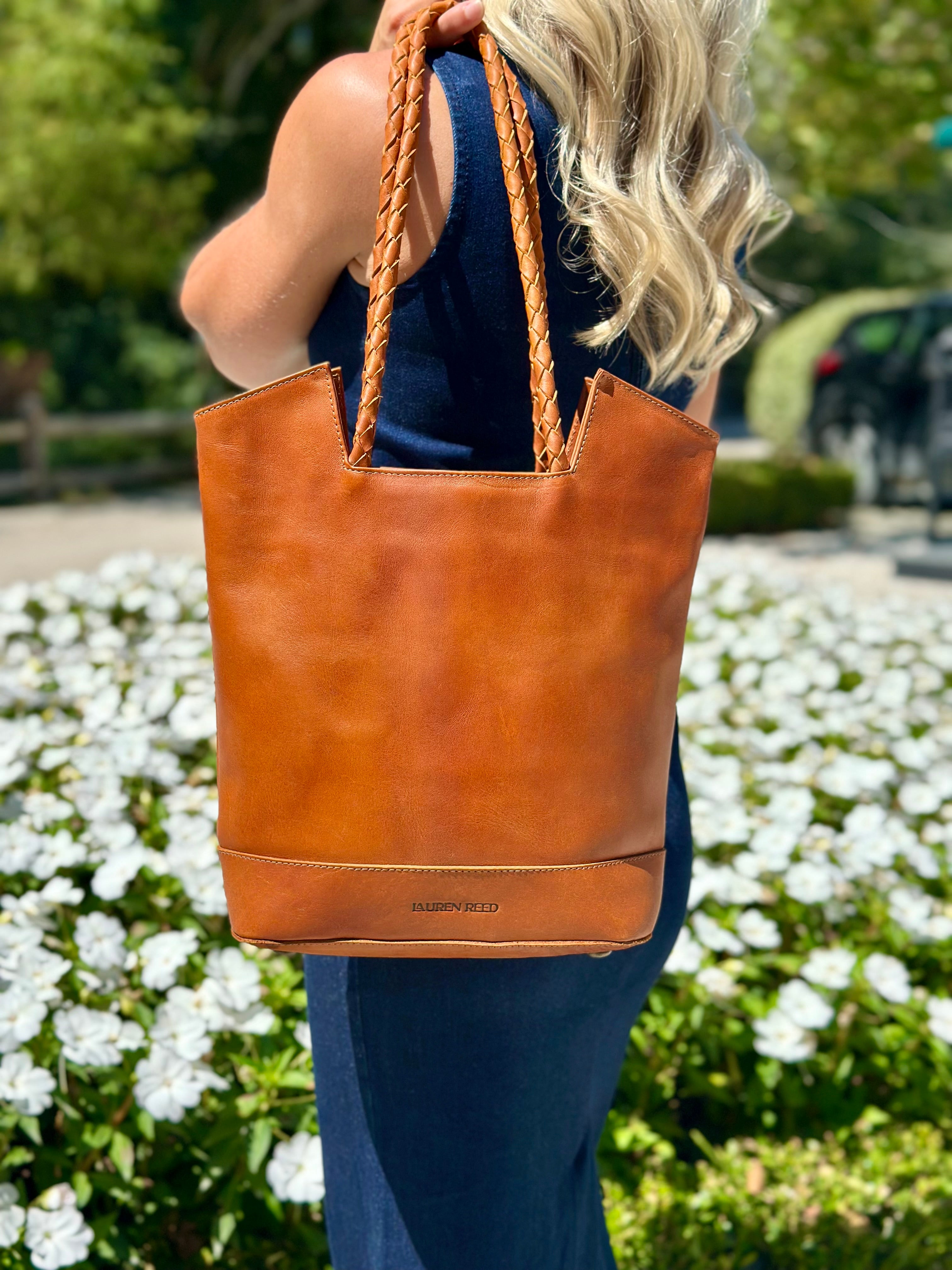 Person holding a tan leather tote bag with braided handles in an outdoor setting.