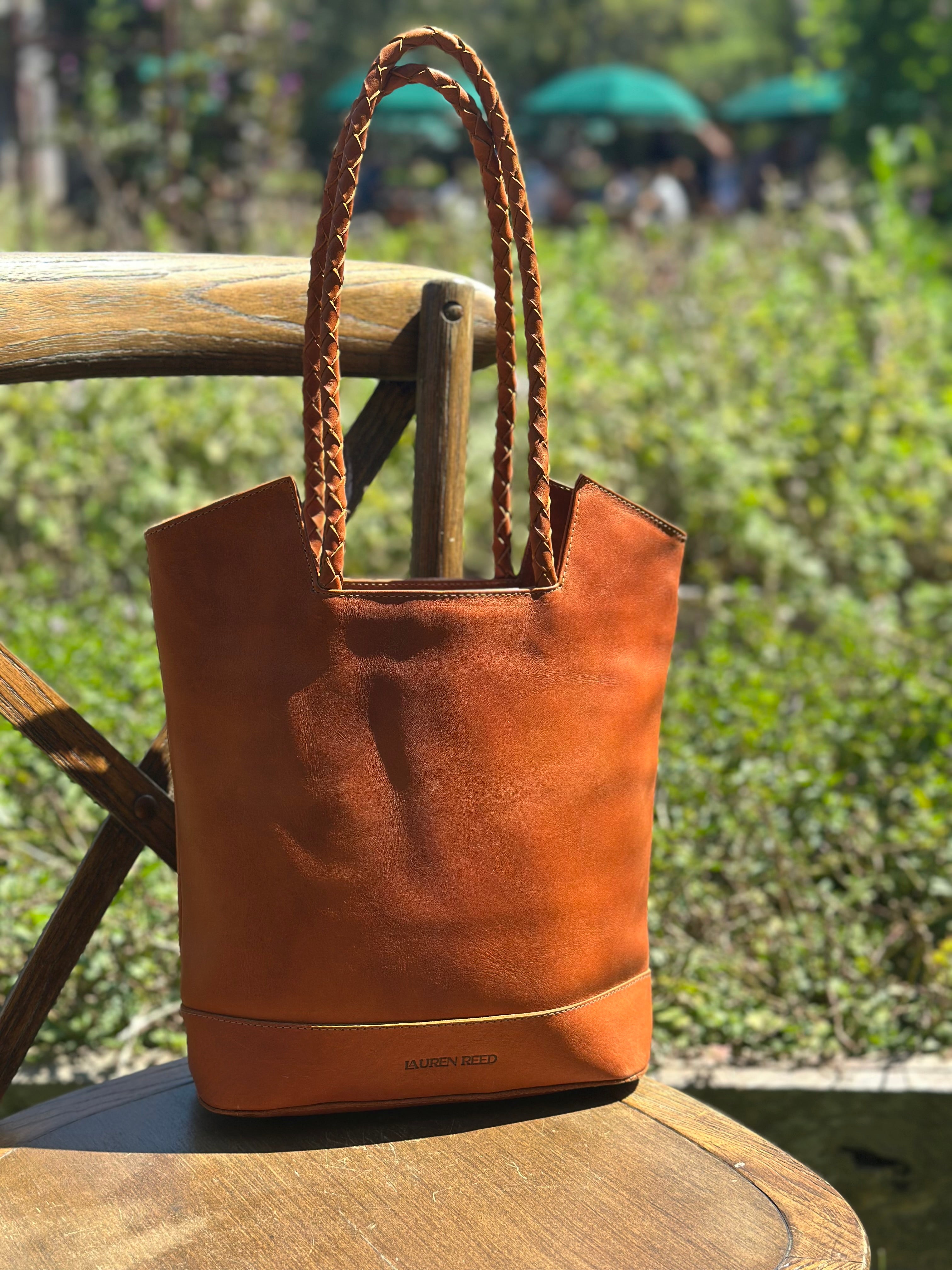 Tan leather tote bag with braided handles on a wooden surface outdoors.