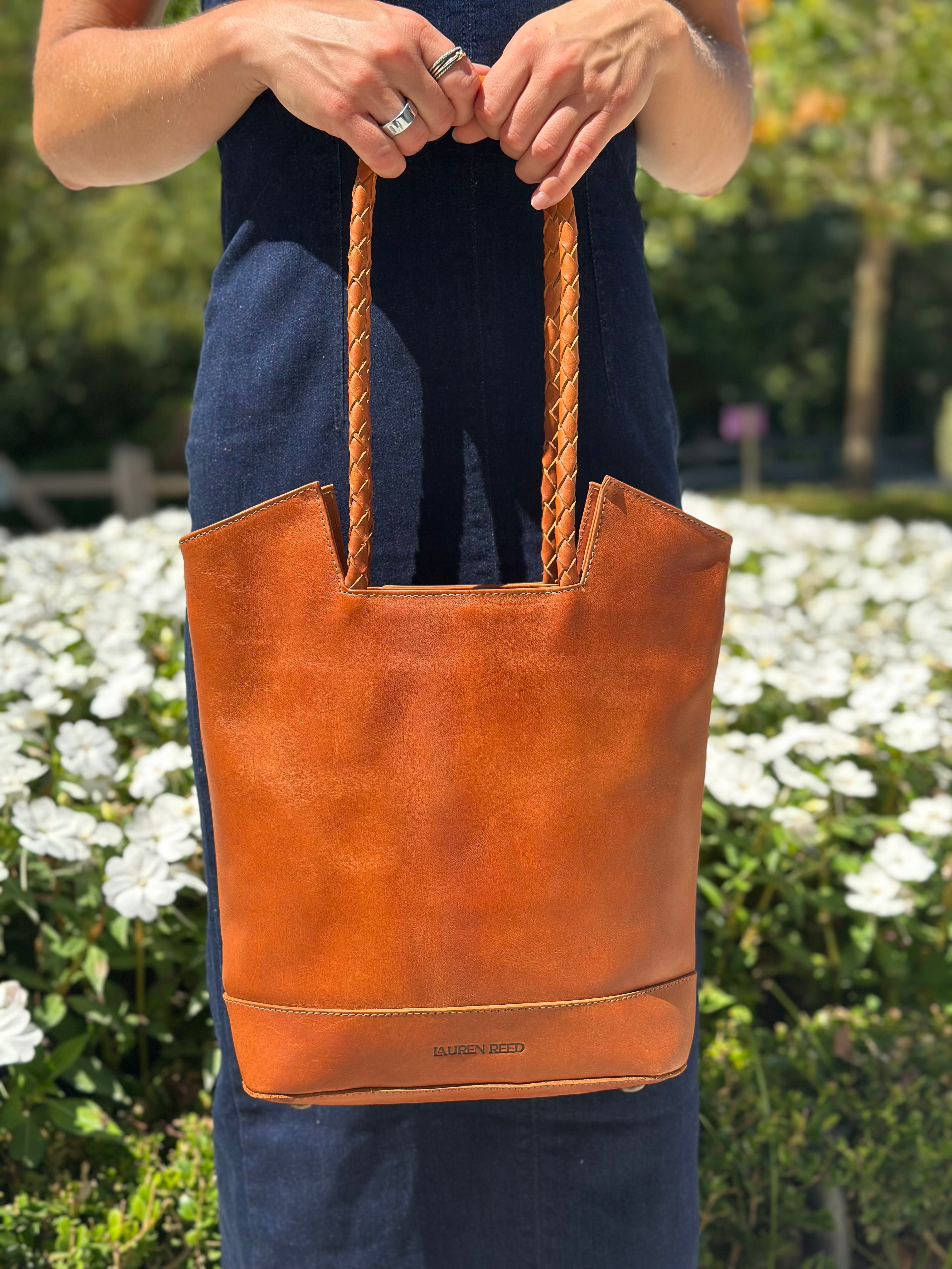 Person holding a tan leather tote bag with braided handles in front of white flowers.