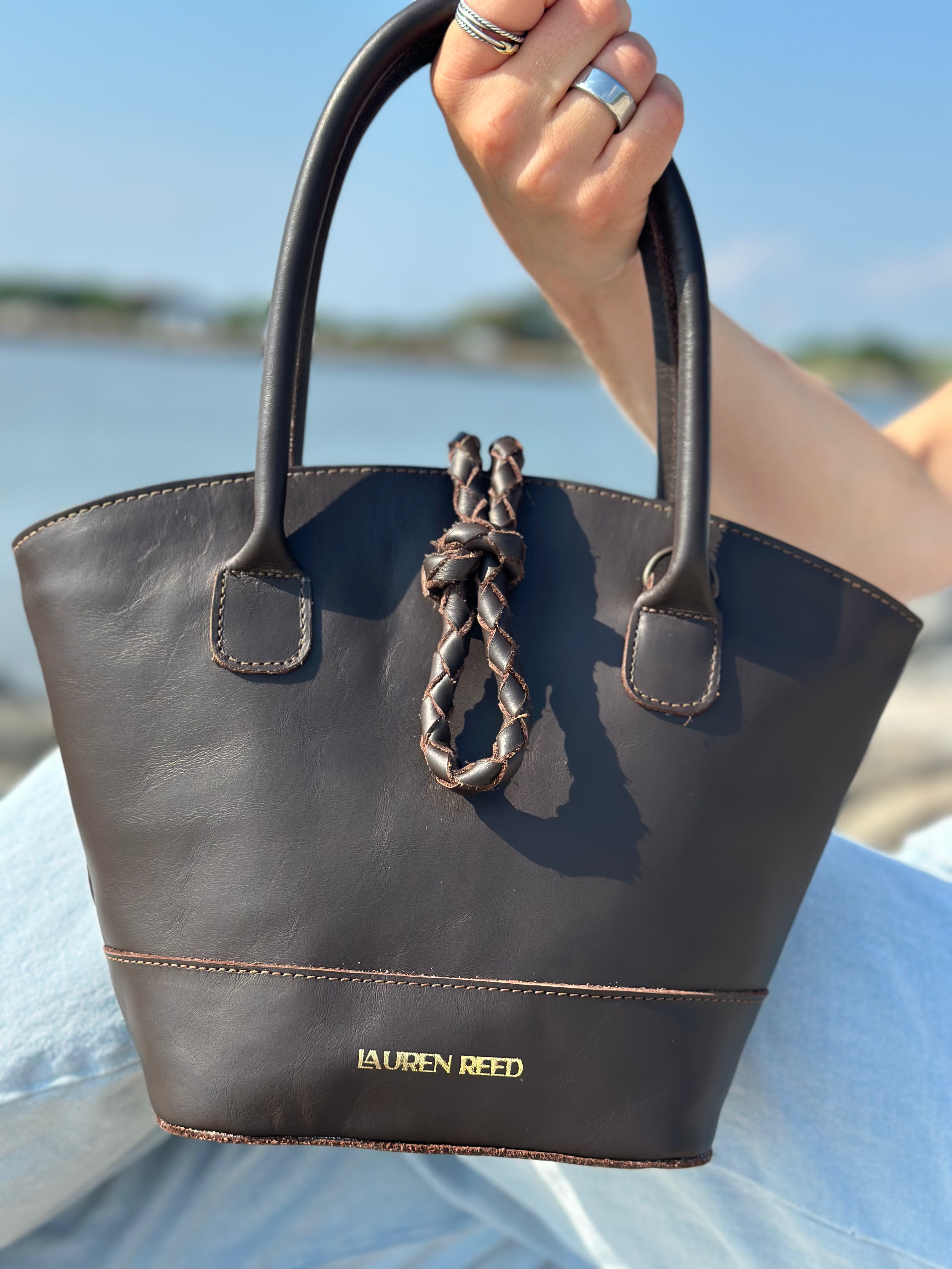 Brown handbag with braided detail held by a person by a body of water.