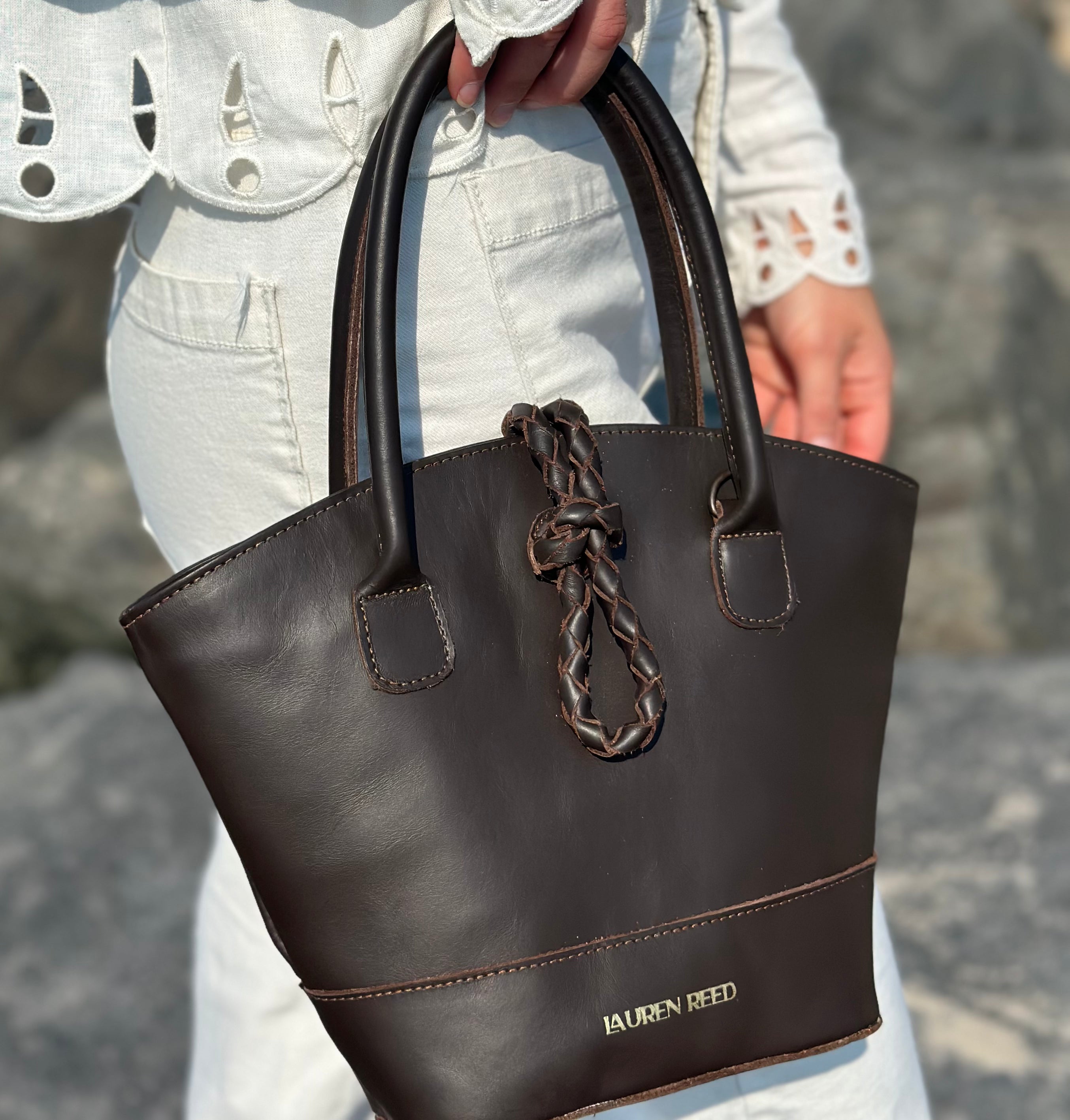 Brown handbag with braided handle held by a person against a blurred background