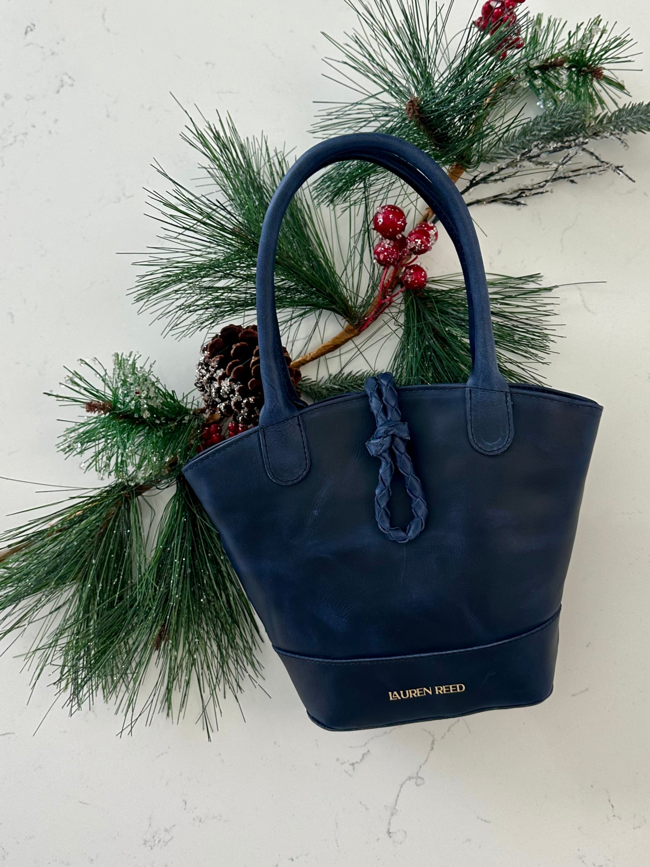 Navy blue handbag with braided closure on a white background with greenery