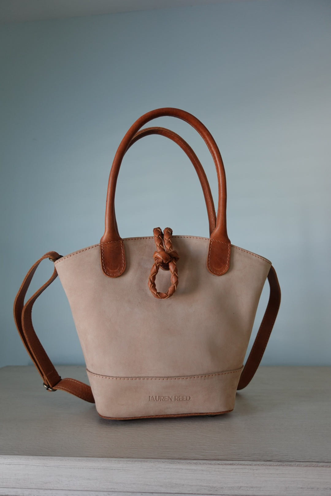 Beige leather handbag with brown handles and trim against a light blue wall. Detachable crossbody purse.