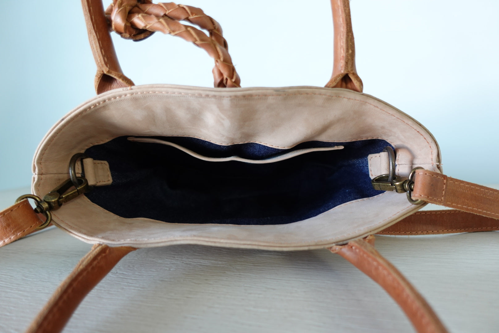 Open beige handbag with denim lining and brown handles and strap on a light blue background
