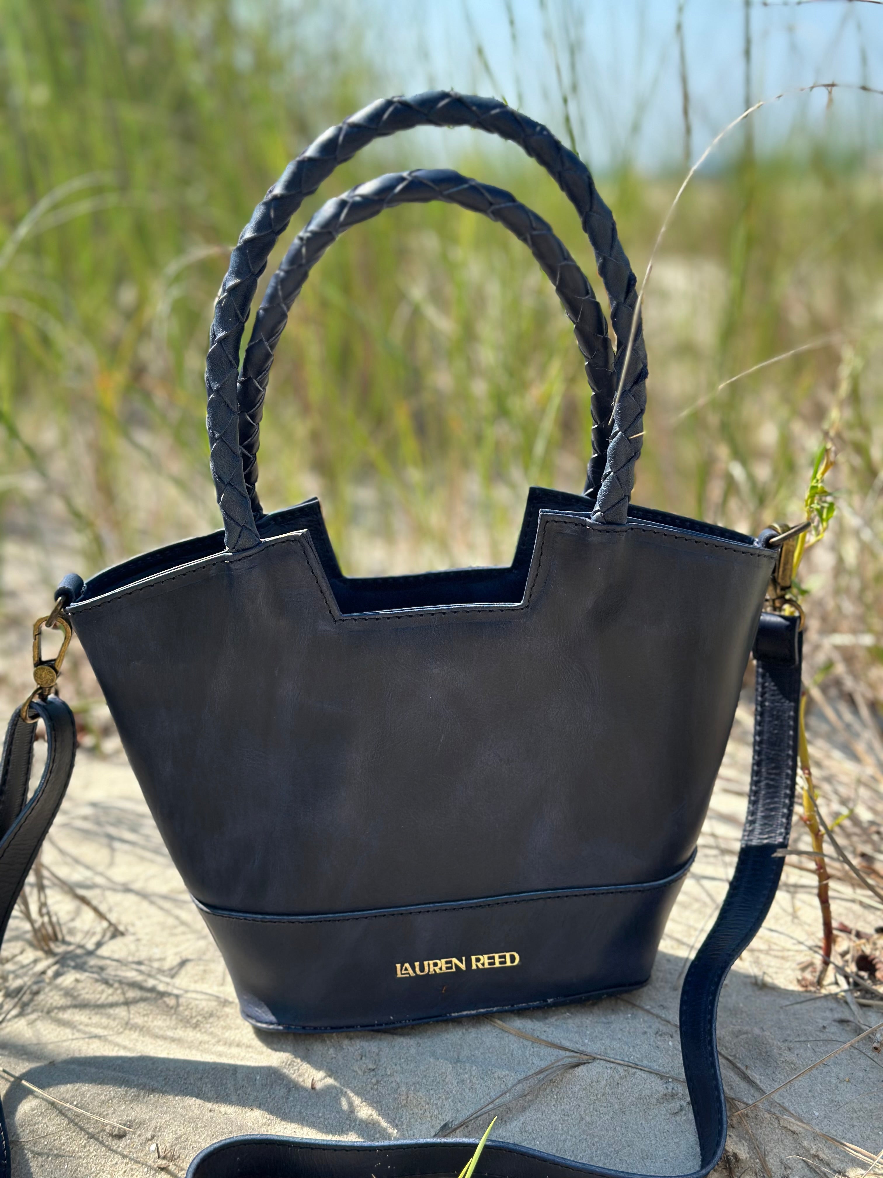 Blue handbag with braided handles on a sandy surface with grass in the background