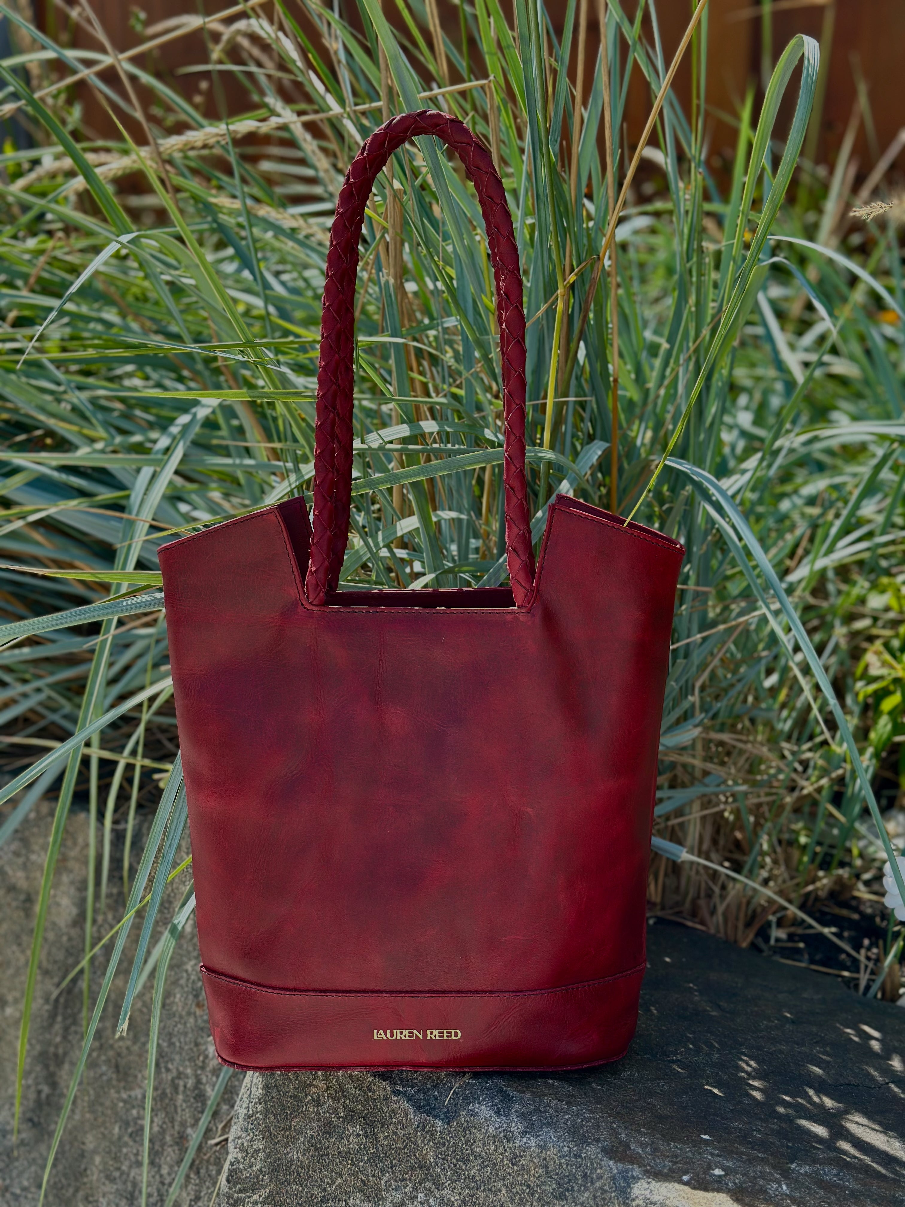 Red handbag with 'Lauren Reed' branding on a stone surface with grass in the background
