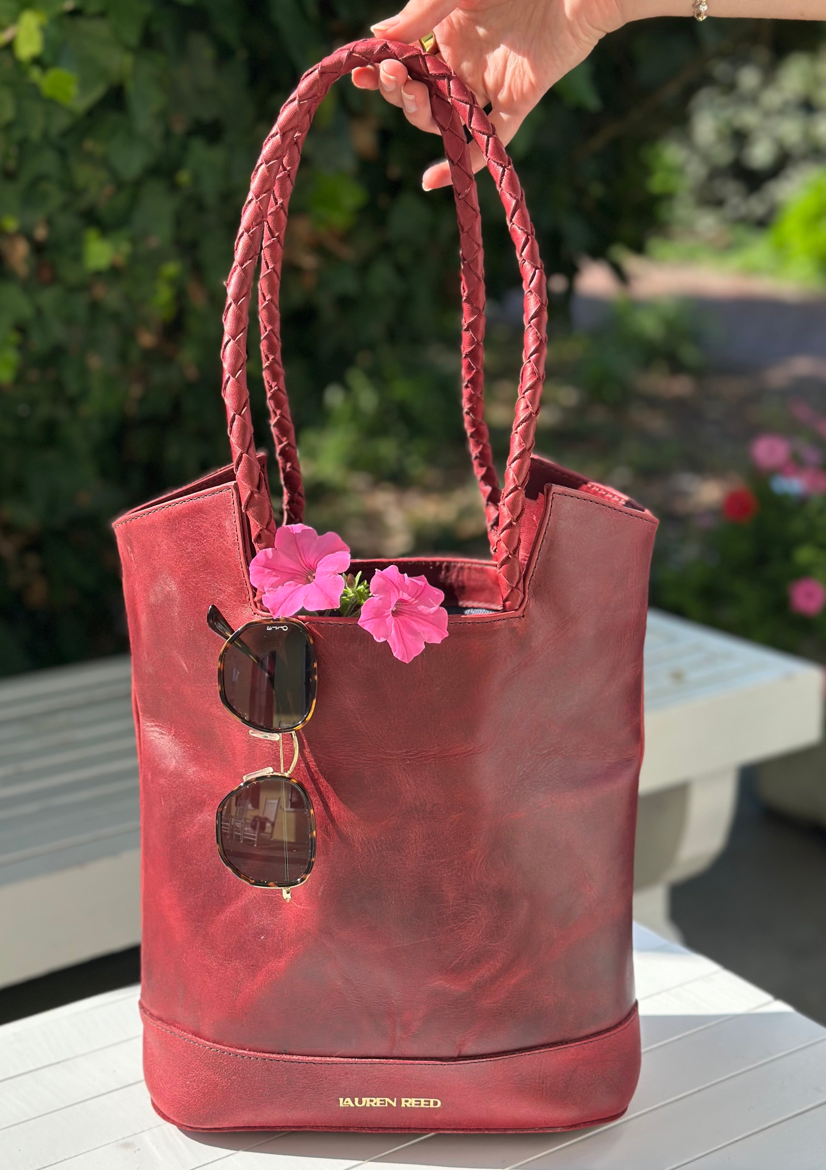 Red handbag with braided handles, sunglasses, and pink flowers on a blurred outdoor background