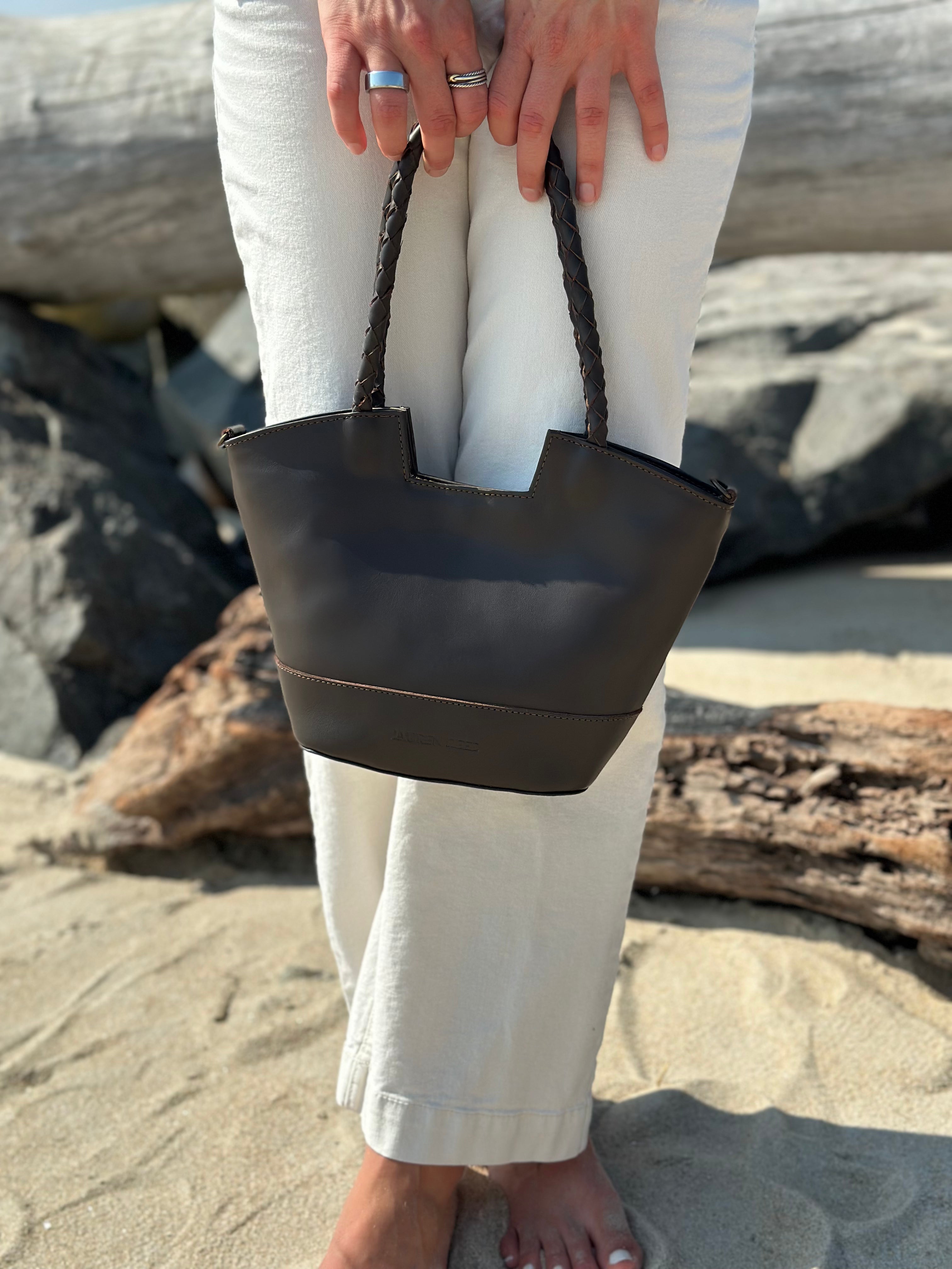 Person holding a brown leather handbag on a beach with rocks and sand in the background.