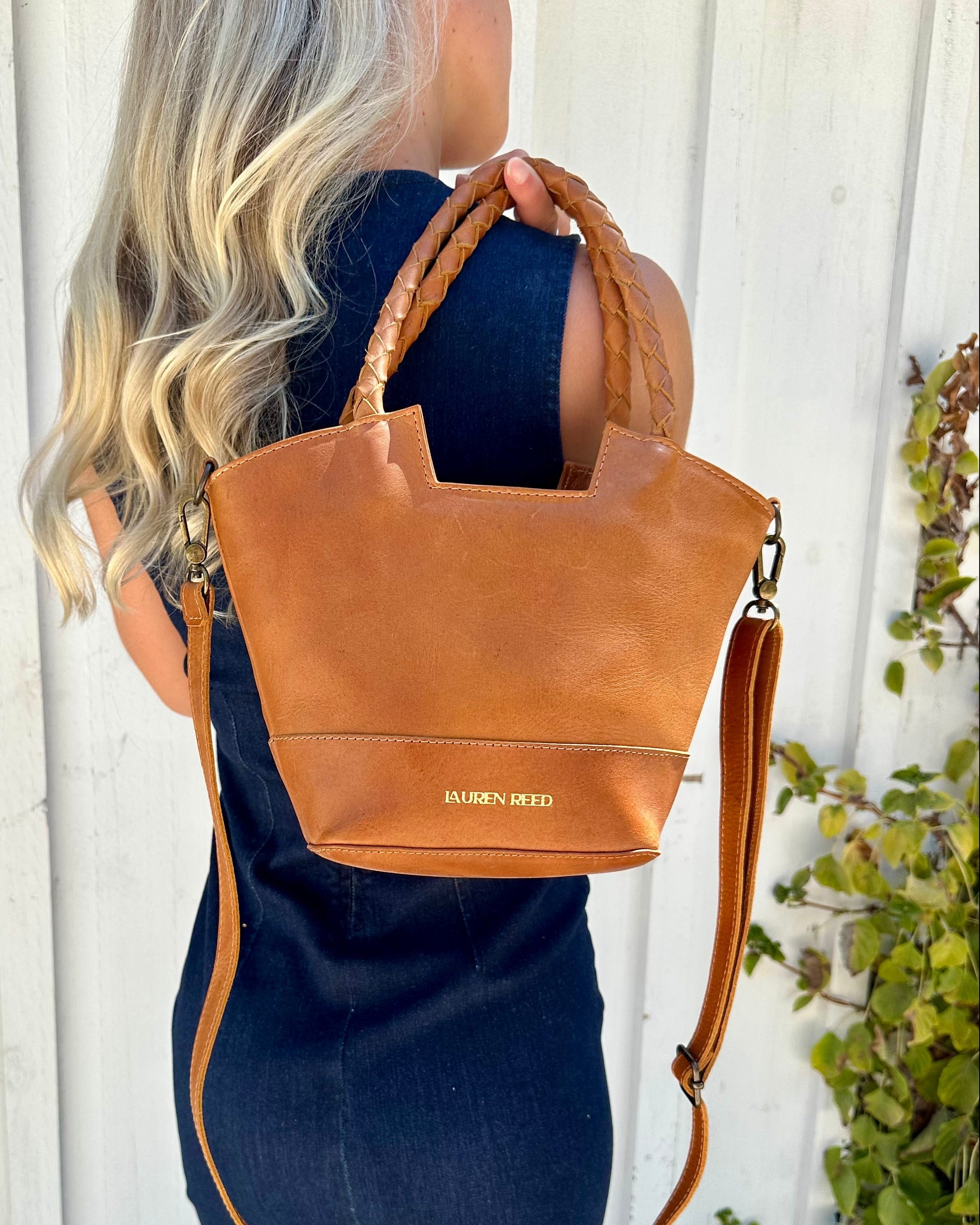 Person holding a tan leather handbag with visible brand name "Lauren Reed" against a white wooden background