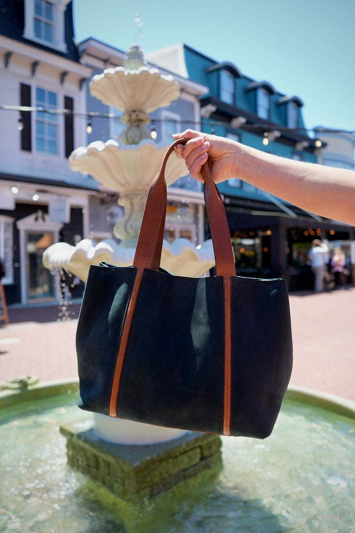 Person holding a navy blue leather tote bag with tan straps in front of a fountain.
