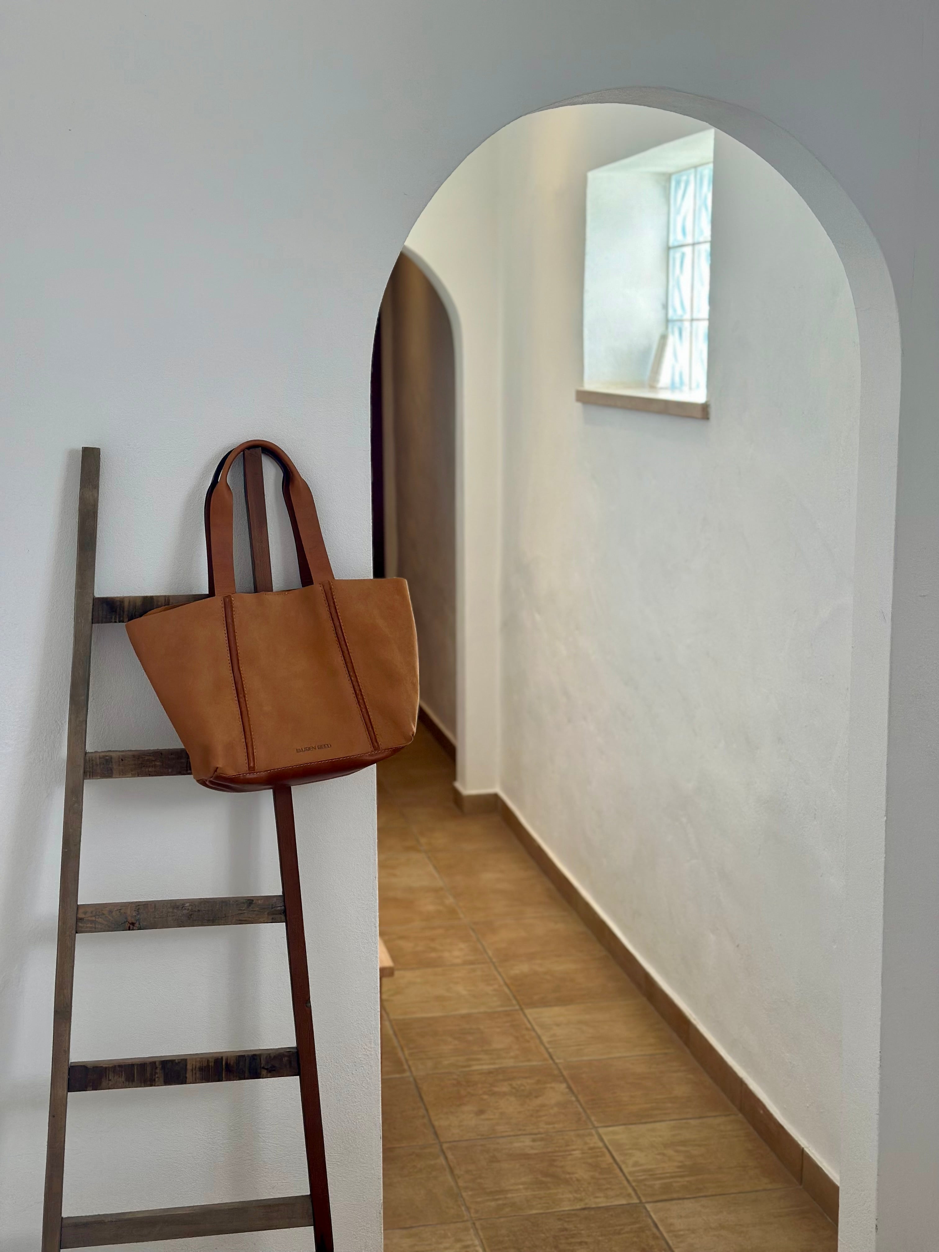 Tan leather bag hanging on a wooden ladder against a white wall.