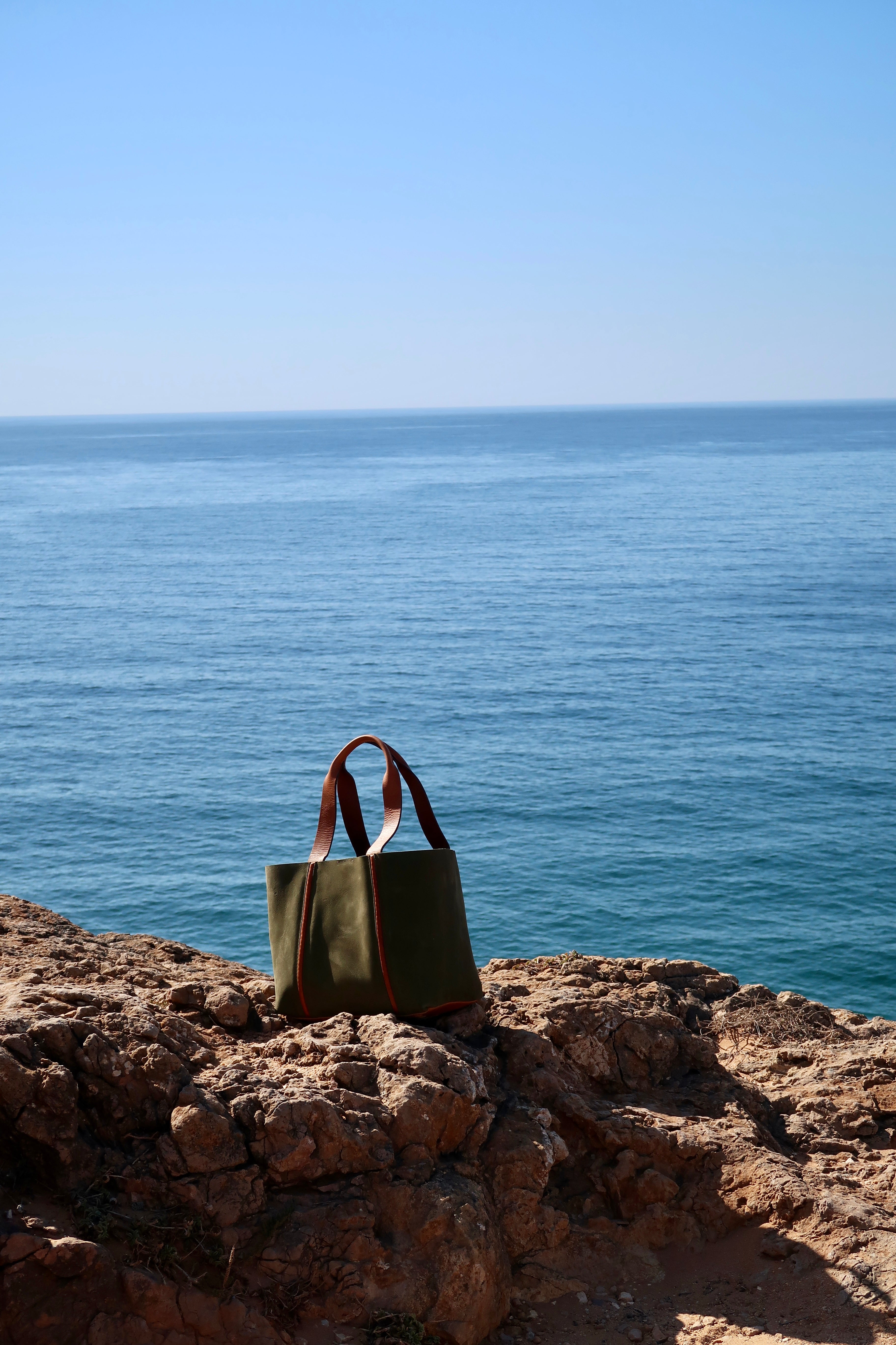Green leather handbag on a rocky outcrop with ocean and blue sky in the background