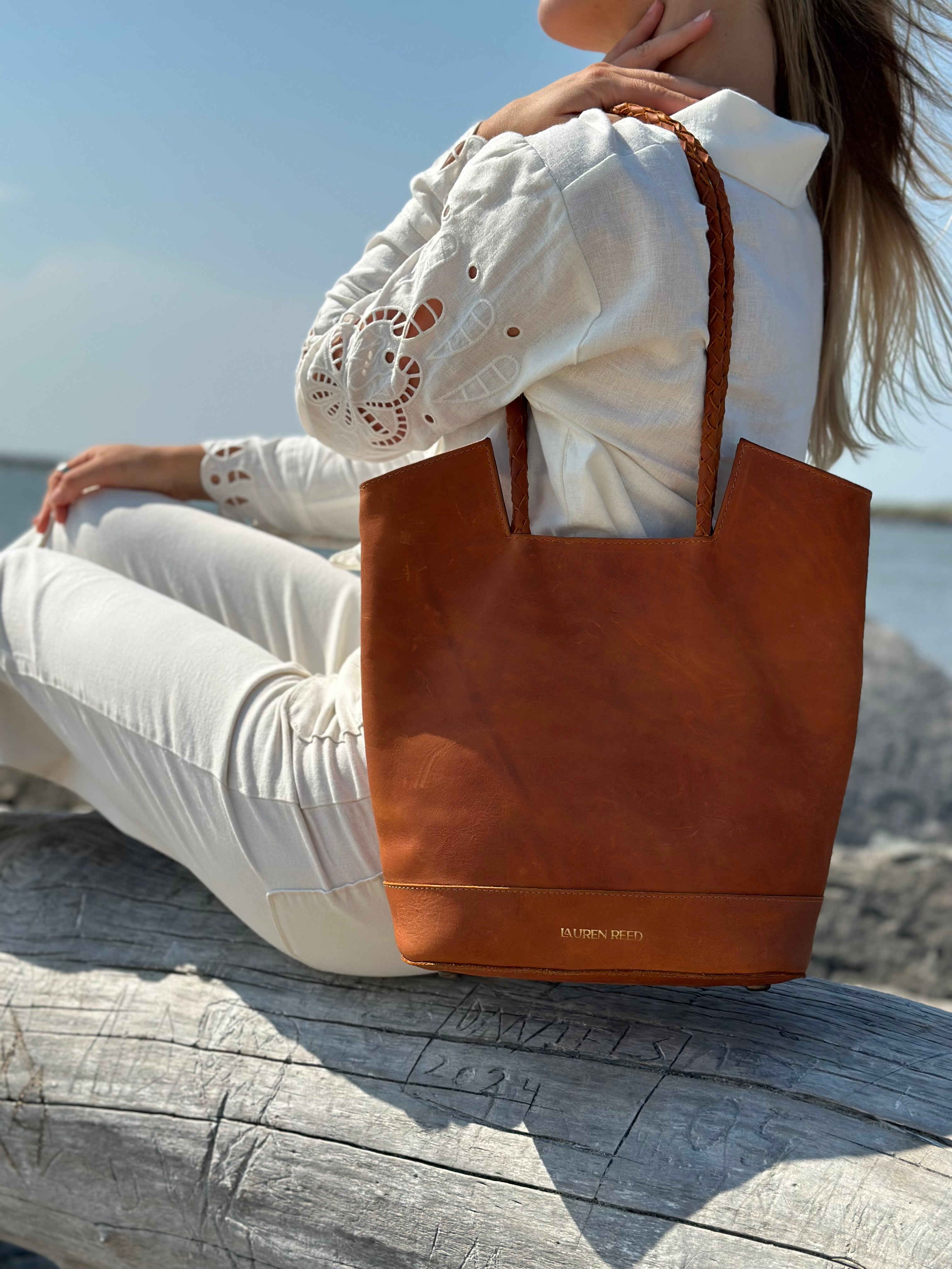 Person sitting on a log by the water with a brown leather tote bag.