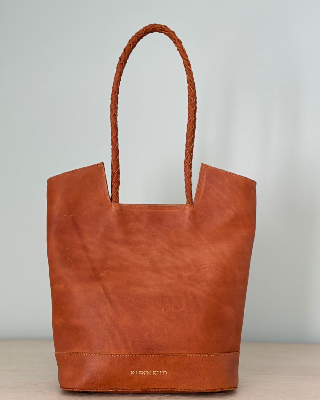 Brown leather tote bag with braided handle on a light gray background