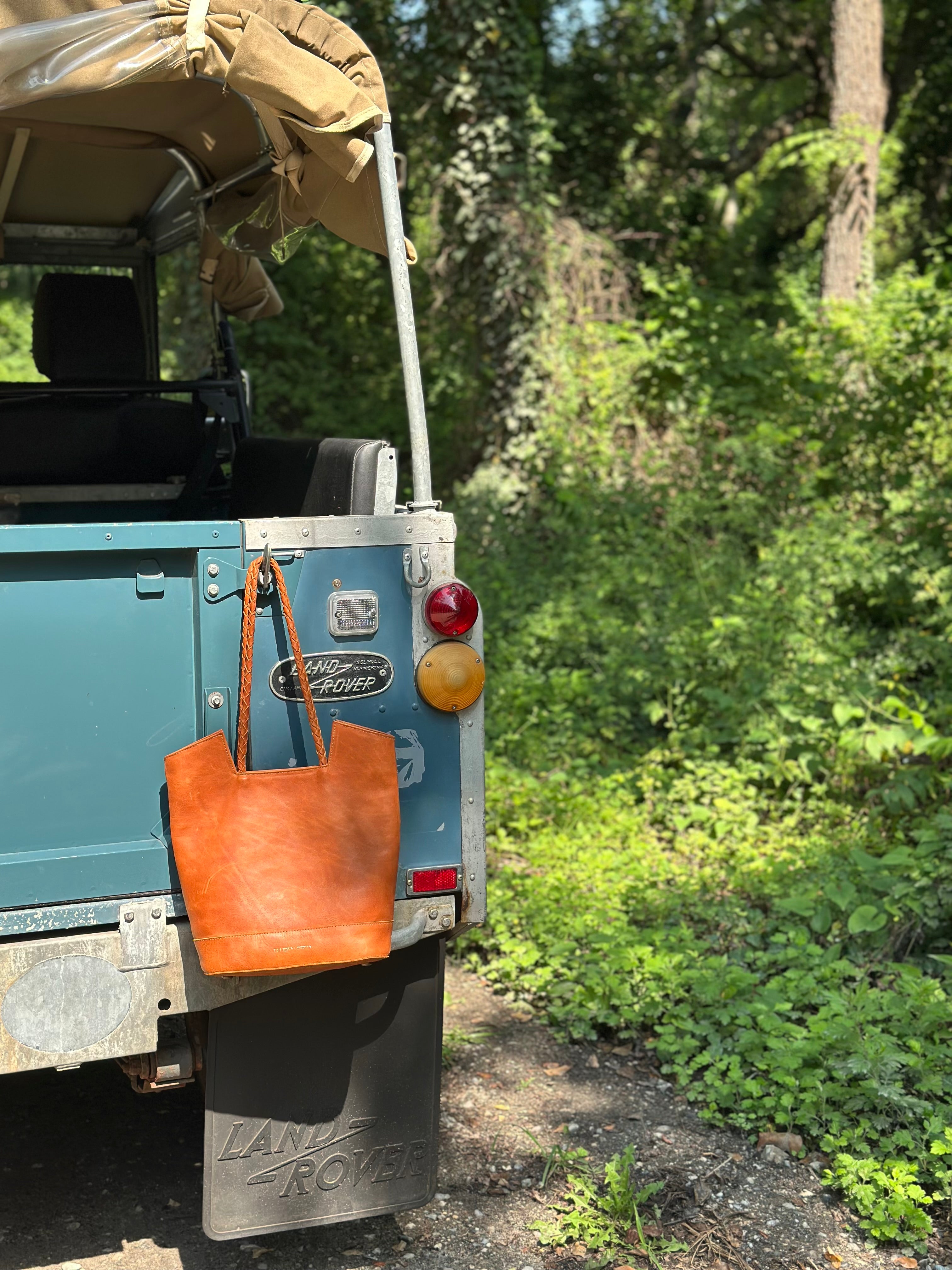 Back of a Land Rover with a brown leather bag hanging off the back, surrounded by greenery.