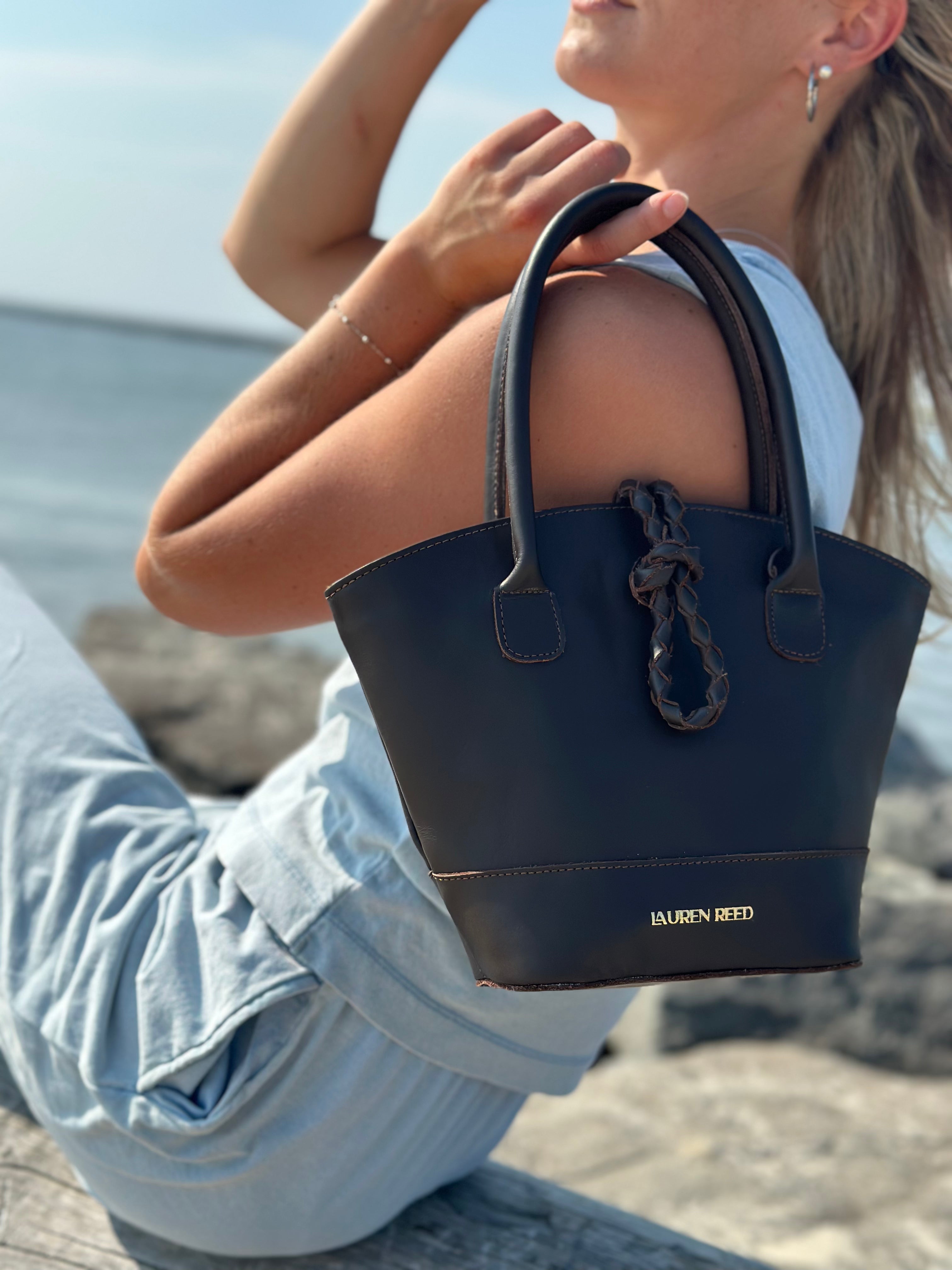 Person holding a brown handbag with 'Lauren Reed' branding by the water.