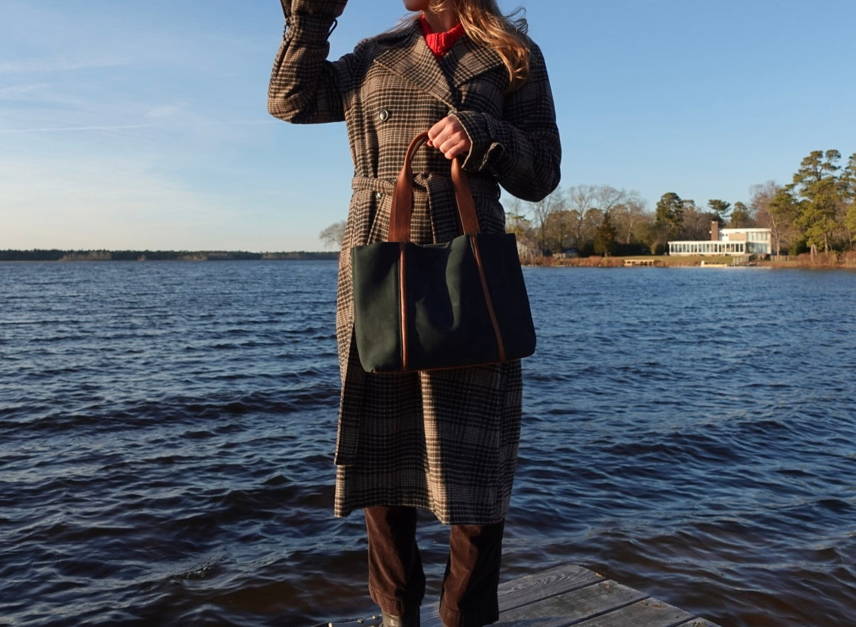 Person standing on a dock by a body of water, wearing a plaid coat and holding a leather handbag.