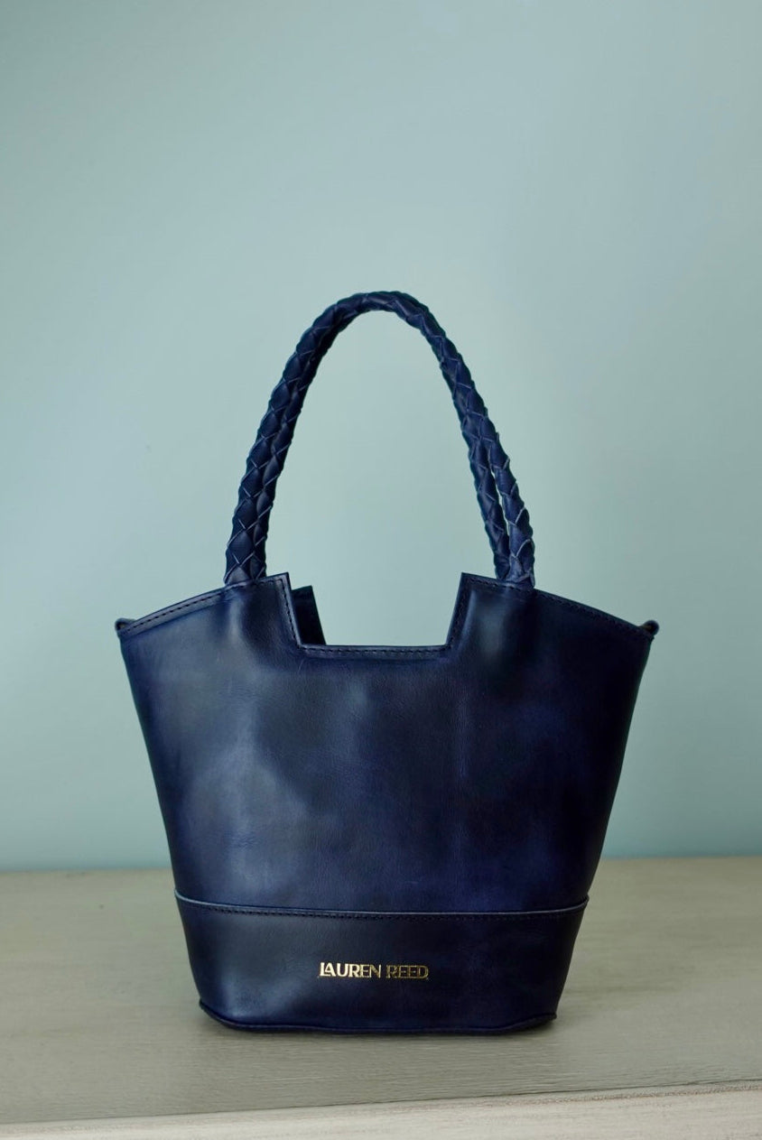Navy blue handbag with braided handle on a light gray surface against a light green background