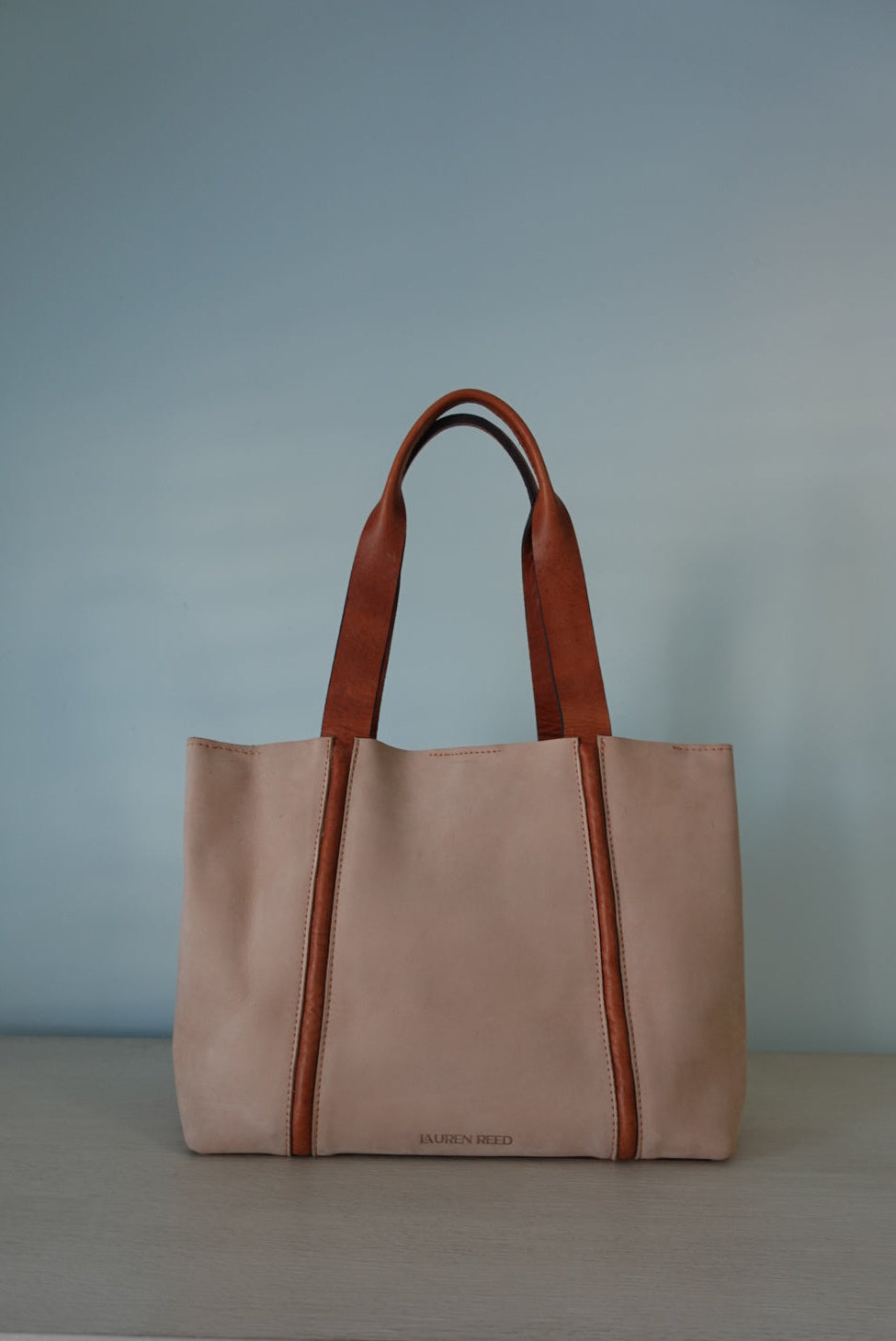 Beige Lauren Reed Co Nubuck Leather tote bag with brown handles on a light blue wall