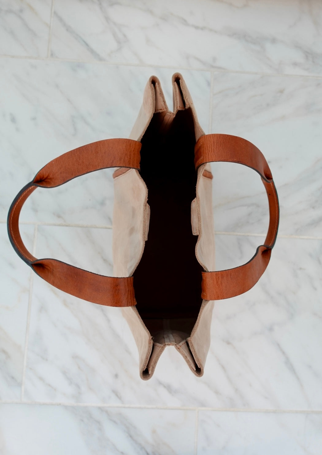 Top-down view of a beige handbag with brown leather straps on a marble surface