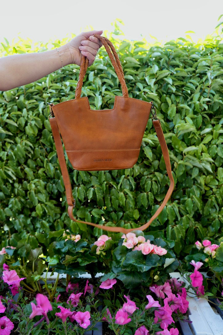 Tan leather summer 2026 handbag held by a person against a green hedge and pink flowers background